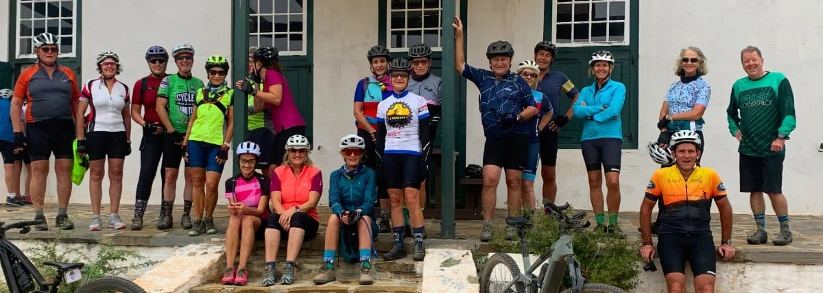 Group of cyclists, mostly women, posing in front of a building, some wearing helmets and cycling gear, with bicycles in front.