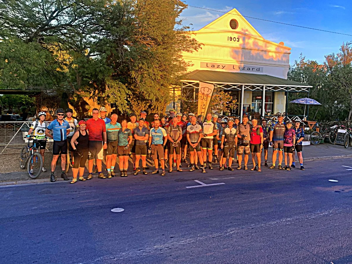 Group of cyclists standing in front of a building with a sign that reads 'Lazy Lizard' during sunset.