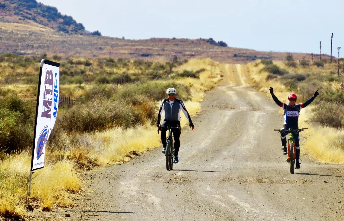 Two cyclists riding on a dirt trail in a desert landscape with dry grass and hills, one raising arms in celebration near a race banner.