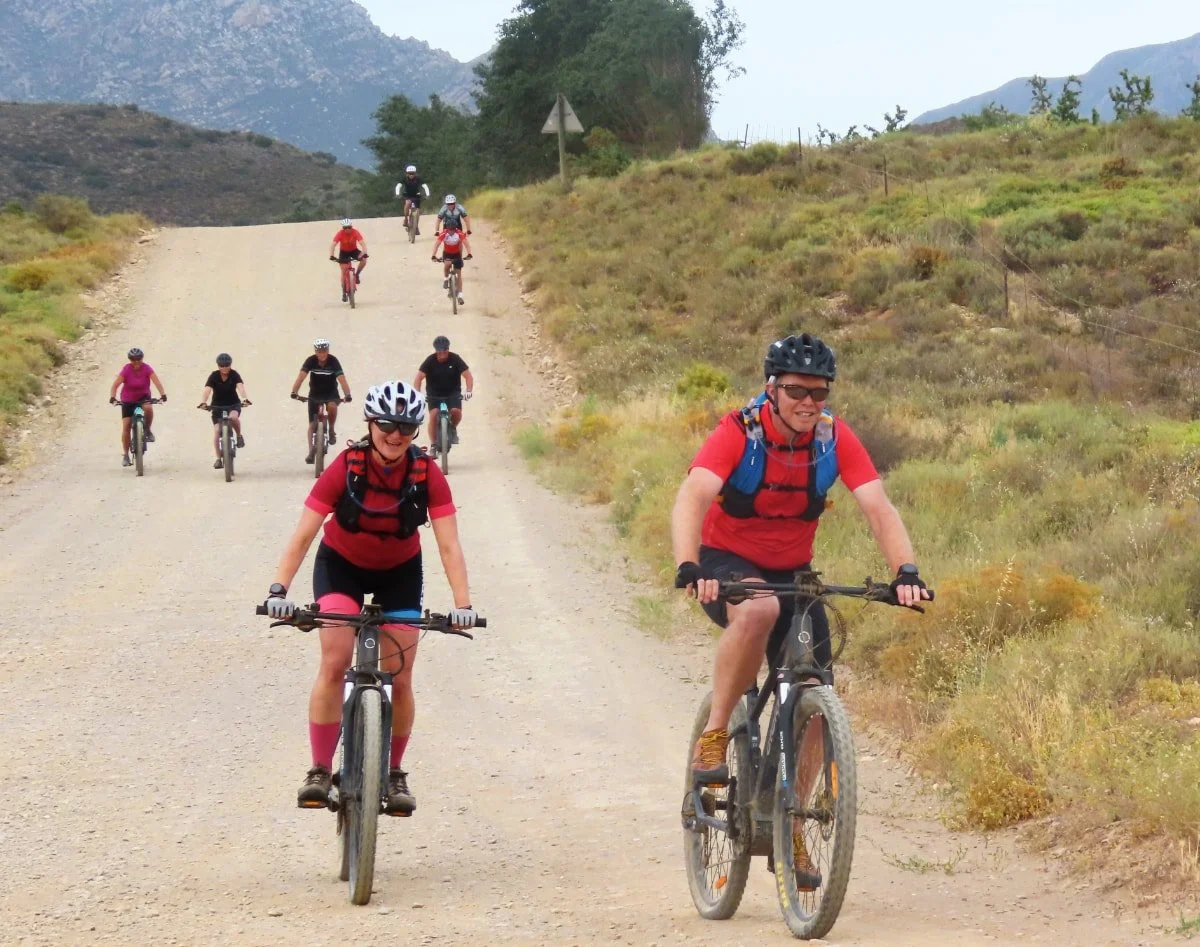 People riding mountain bikes on a dirt trail in a rural landscape with hills and mountains in the background.