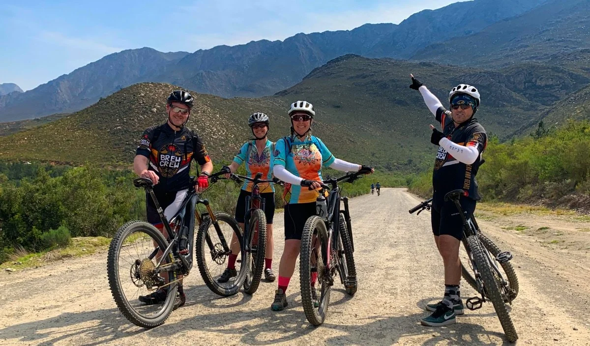Four people with mountain bikes on a dirt trail in a mountainous area, wearing helmets and colorful cycling jerseys, smiling and posing for a photo.