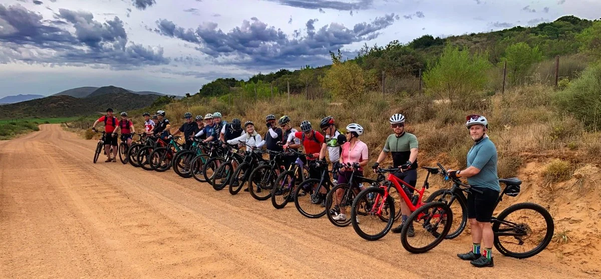 Group of cyclists standing with their bikes on a dirt road in a hilly outdoor area.