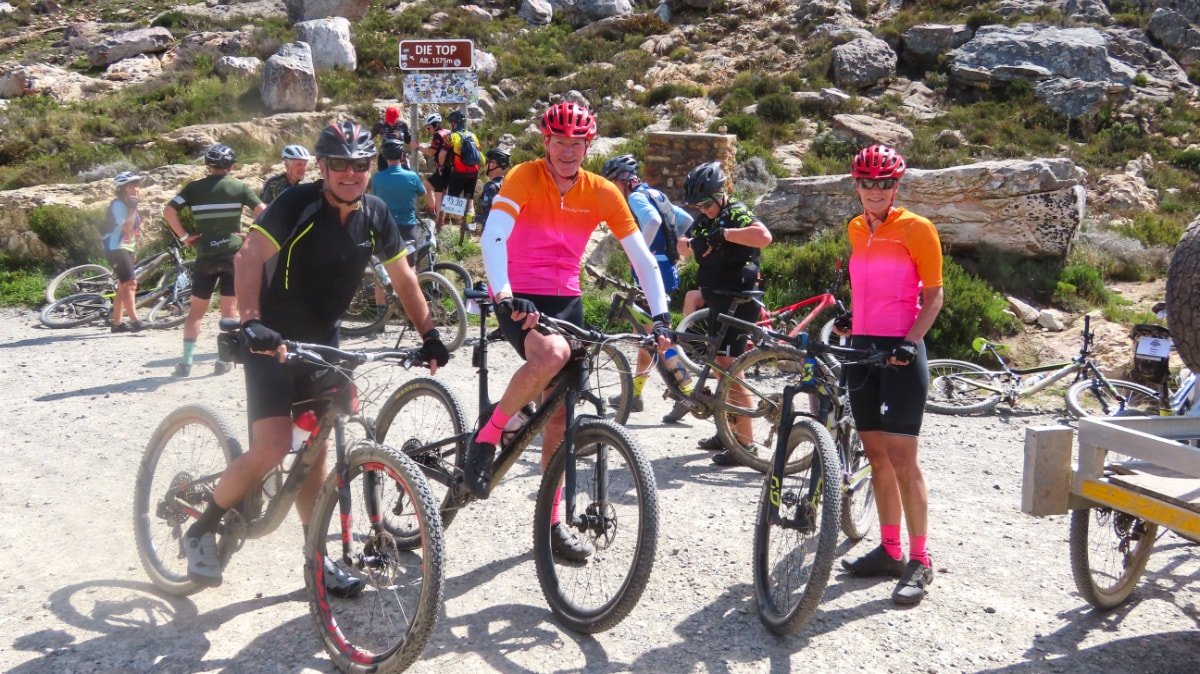 Group of cyclists with mountain bikes at a high-altitude location, indicated by a sign that reads 'DIE TOP 1757 M', in a rocky, arid landscape.