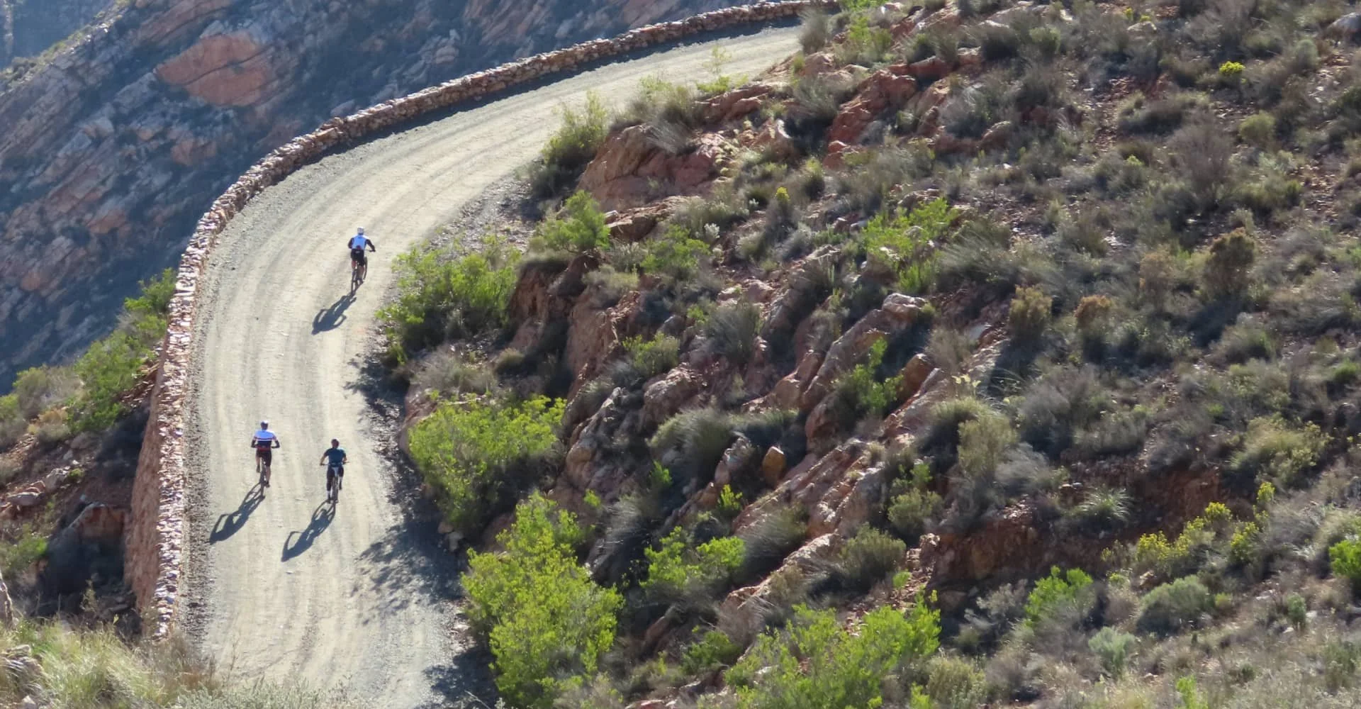 Four cyclists riding up a dirt mountain road with a stone barrier on the left and rocky terrain with sparse vegetation on the right.