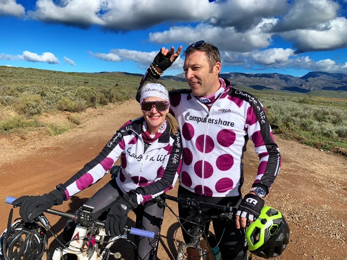 Two cyclists, a woman and a man, stand on a dirt trail with mountains and a blue sky with clouds in the background. They are wearing matching pink and white polka dot jerseys and black gloves, with the woman wearing sunglasses and a headband, and the