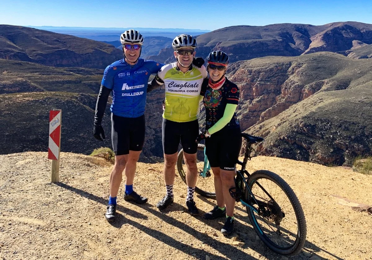 Three cyclists in colorful gear and helmets standing together on a mountain trail with scenic rocky hills in the background.