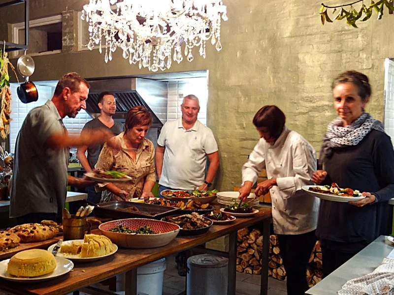 Six people serving themselves food at a dinner buffet table, with a chandelier hanging above and a brick wall in the background.