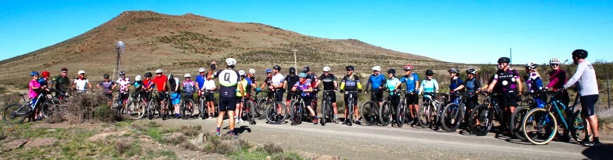 Group of people with bicycles gathered outdoors on a dirt road in front of a hill under a clear blue sky.