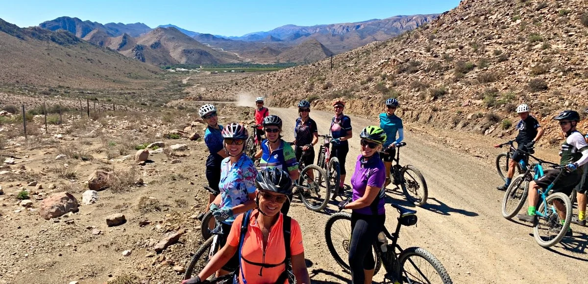 Group of women on mountain bikes taking a break on a dirt trail in a desert landscape with mountains in the background.
