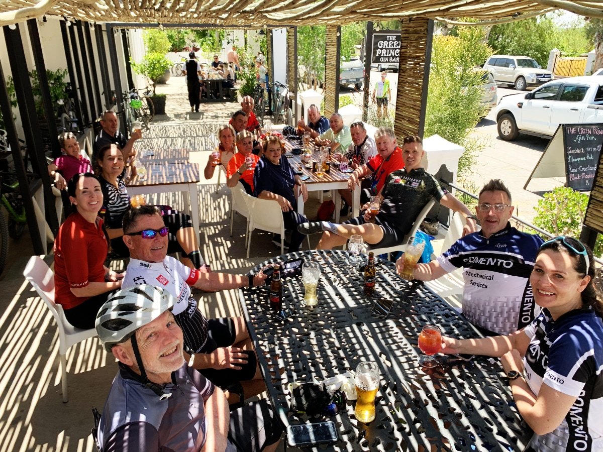 Group of people enjoying drinks at an outdoor patio of a cafe or restaurant, with some wearing cycling jerseys, bicycles parked nearby, and a sign for The Green Prince Gin Bar.