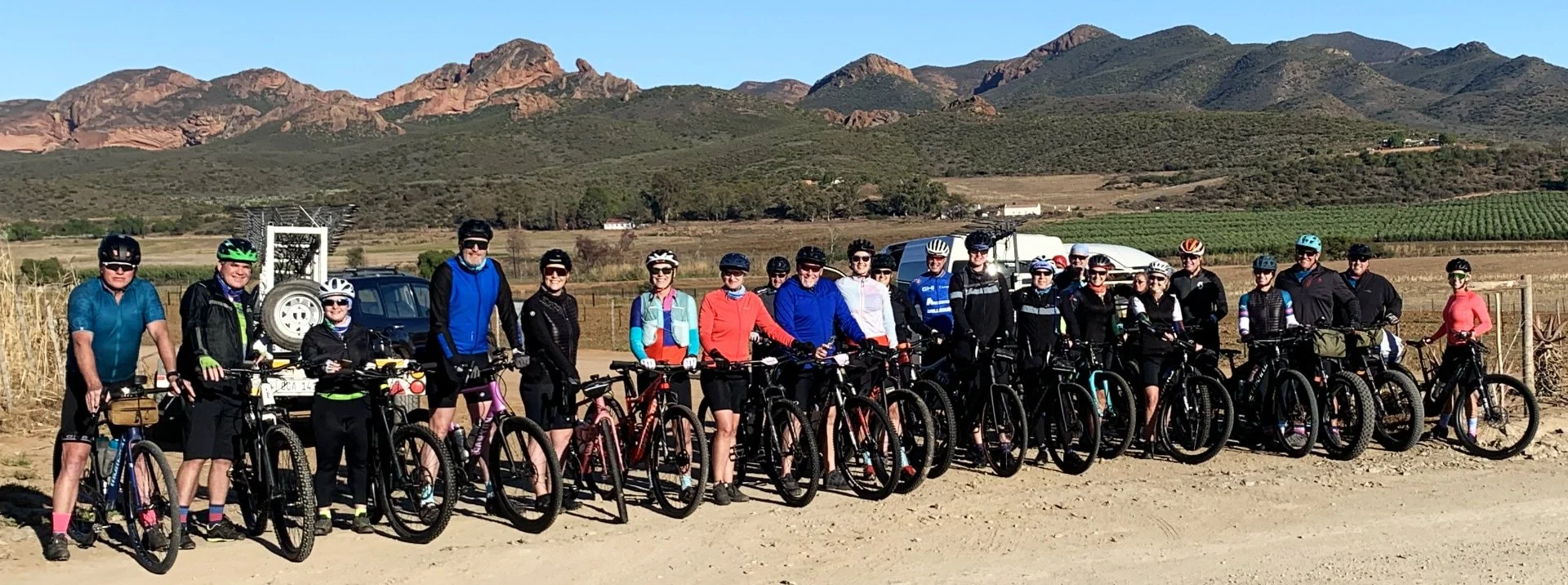 Group of people with bicycles posing together outdoors in a rural area with mountains in the background.