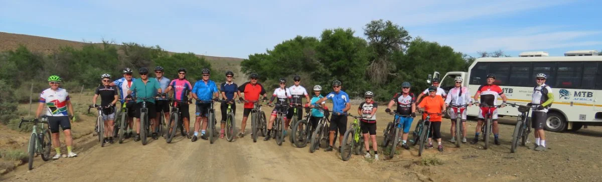 Group of cyclists posing with their bikes on a dirt trail, with a bus and green trees in the background.