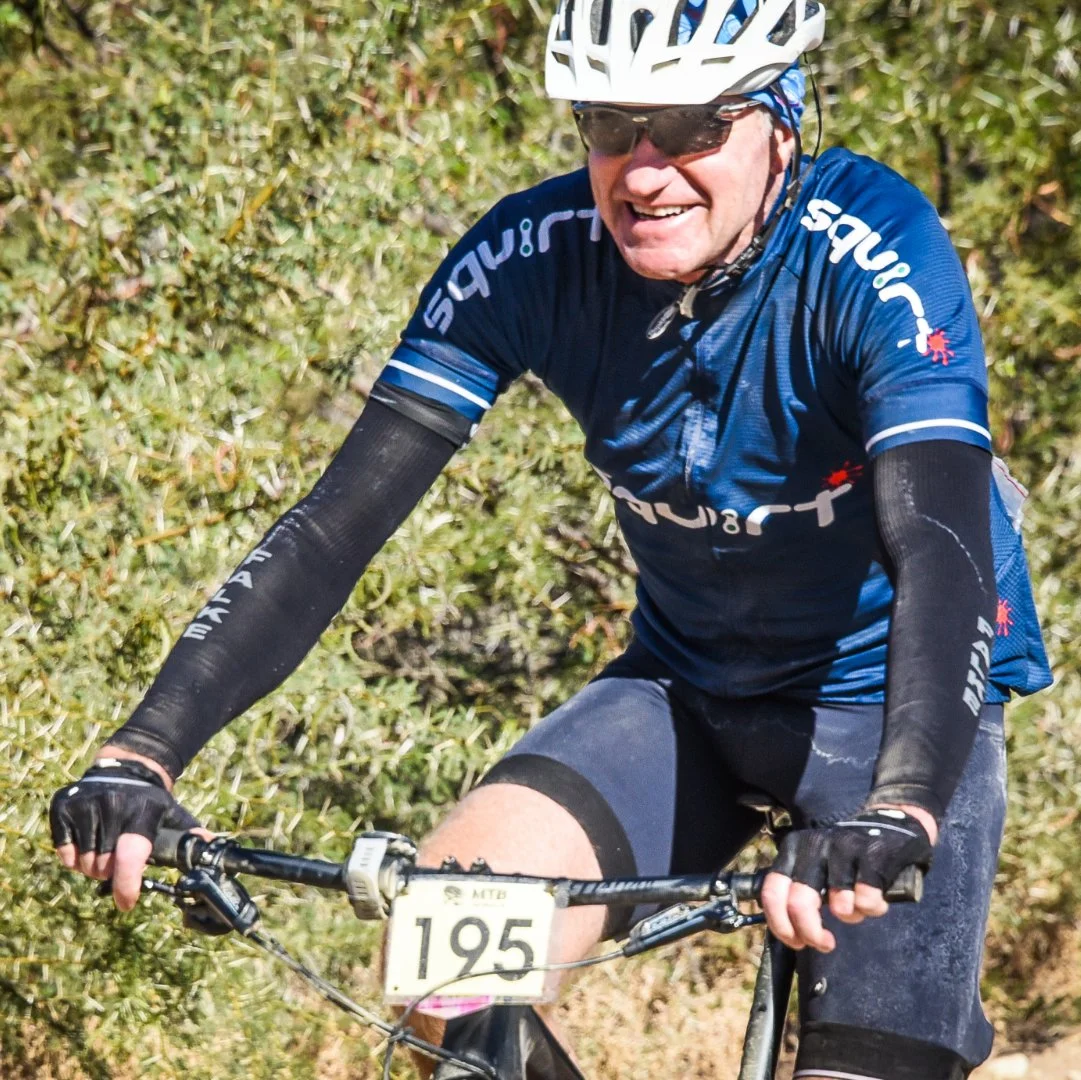 A man riding a mountain bike on a trail surrounded by bushes. He is wearing a blue cycling jersey, black gloves, a white helmet, and sunglasses, smiling as he pedals.