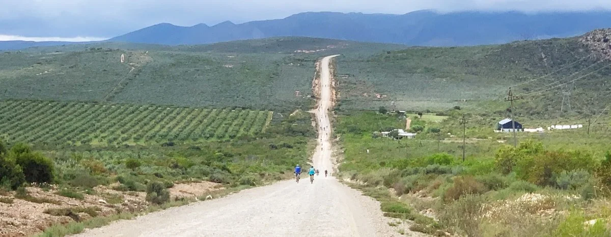 People walking along a long, dusty dirt road through a rural landscape with green farms, telephone poles, and mountains in the distance.