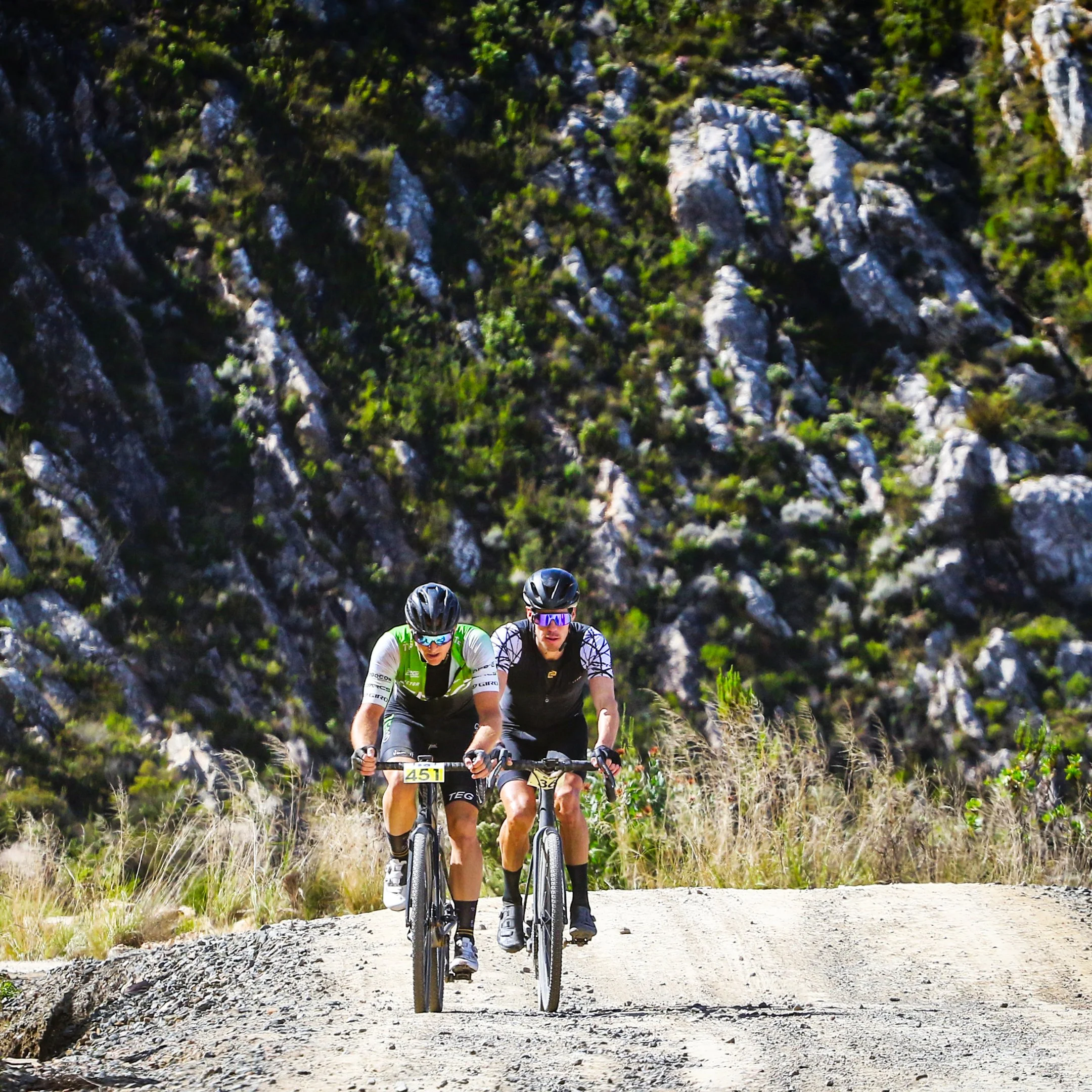 Two cyclists riding mountain bikes on a dirt trail with rocky hills and green vegetation in the background.