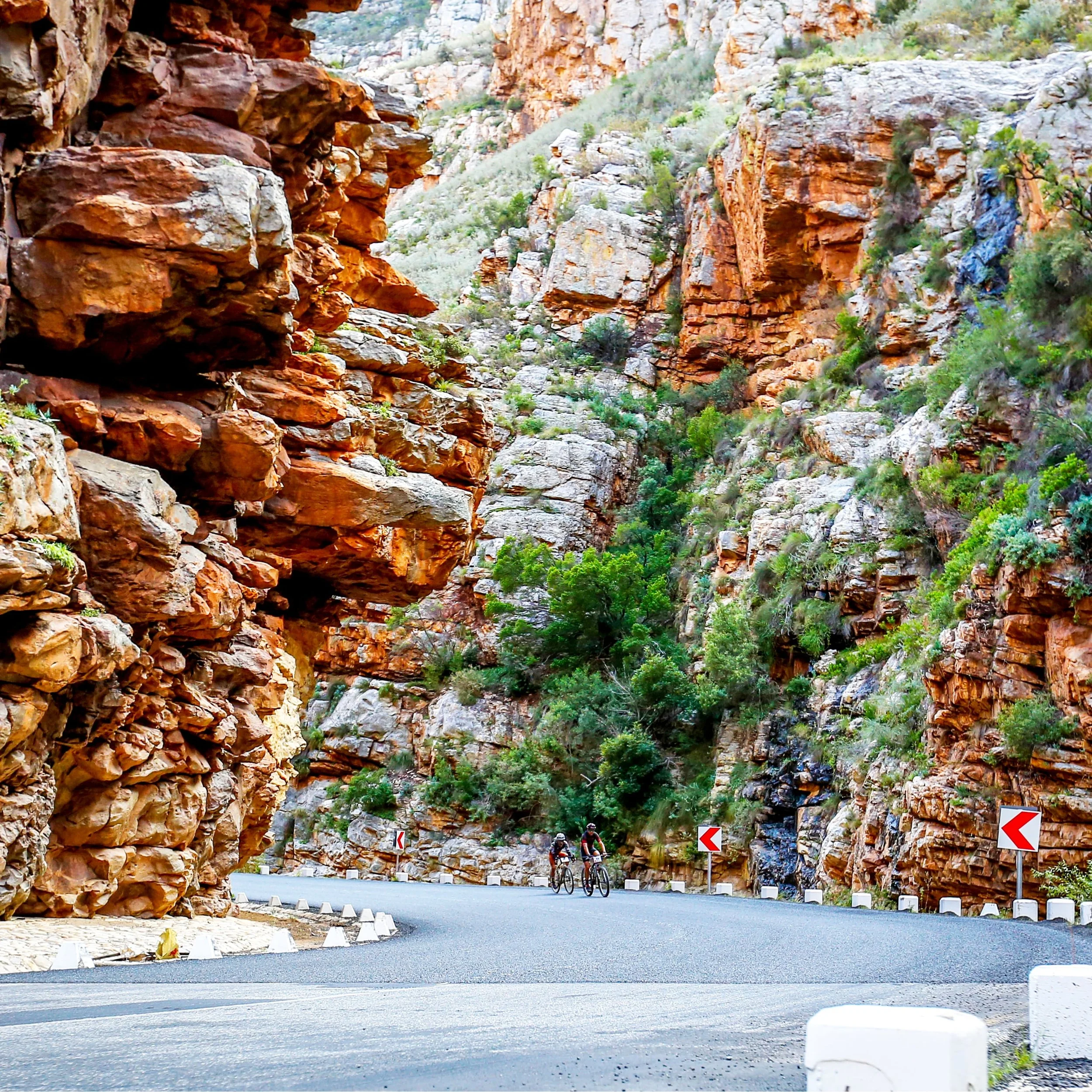 Two cyclists riding on a winding mountain road surrounded by tall red and orange jagged rocks and sparse green vegetation.