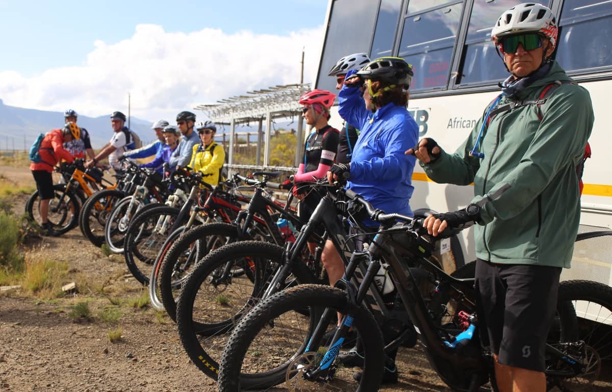 Group of cyclists with helmets and athletic clothing, standing with their bikes lined up on a dirt trail near a bus, in an outdoor setting with mountains and cloudy sky in the background.