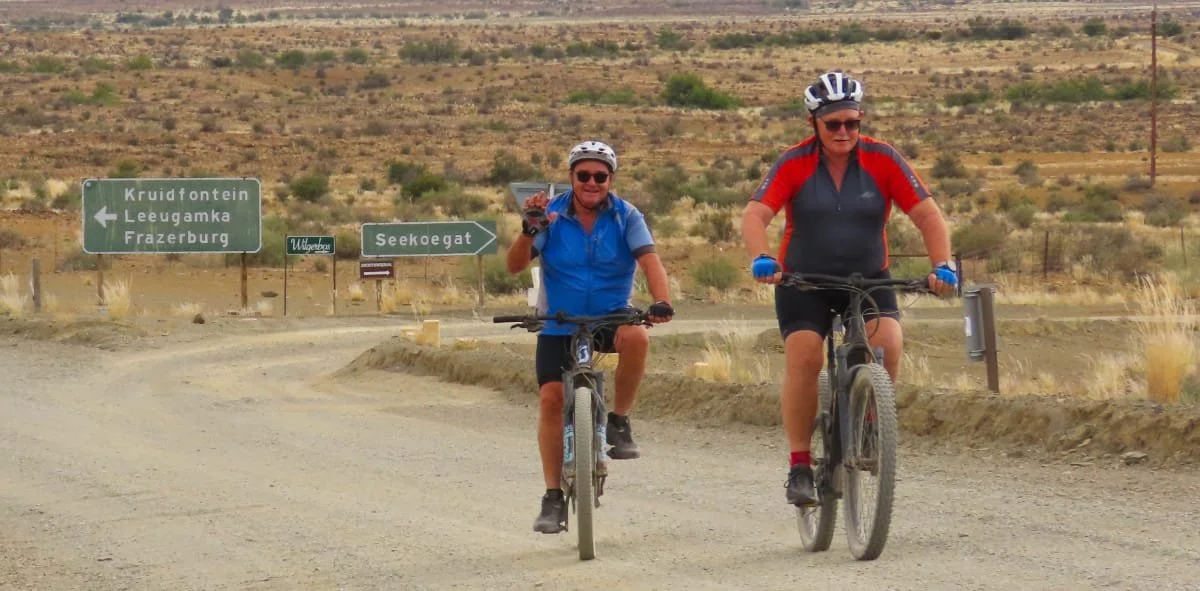 Two people riding mountain bikes on a dirt trail in a desert landscape, with road signs in the background indicating nearby locations.