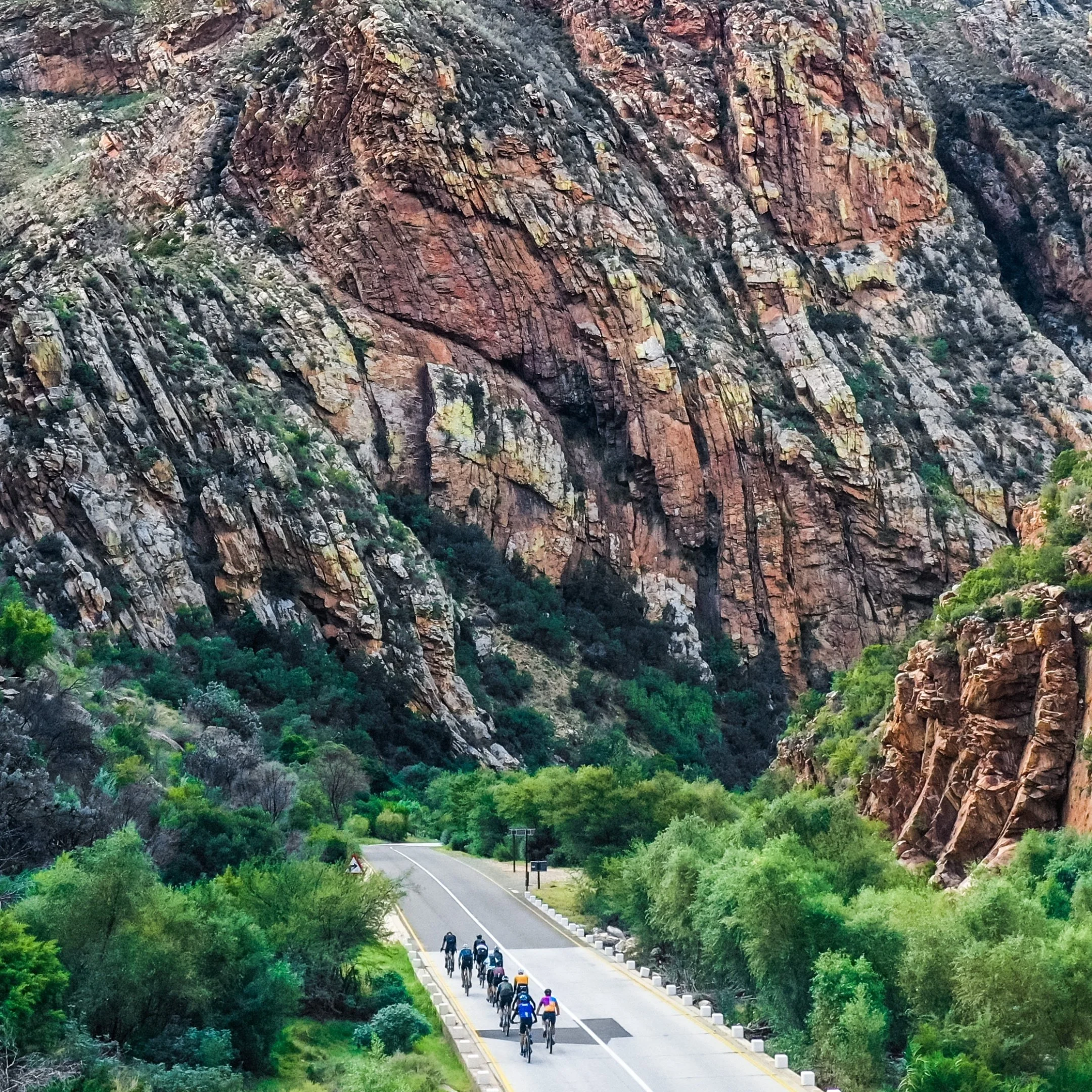 A group of cyclists riding on a road through a canyon surrounded by steep, colorful rocky cliffs and green vegetation.