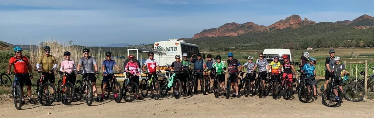Group of 16 mountain bikers posing together outdoors with bicycles, mountains in the background, and a bus behind them.