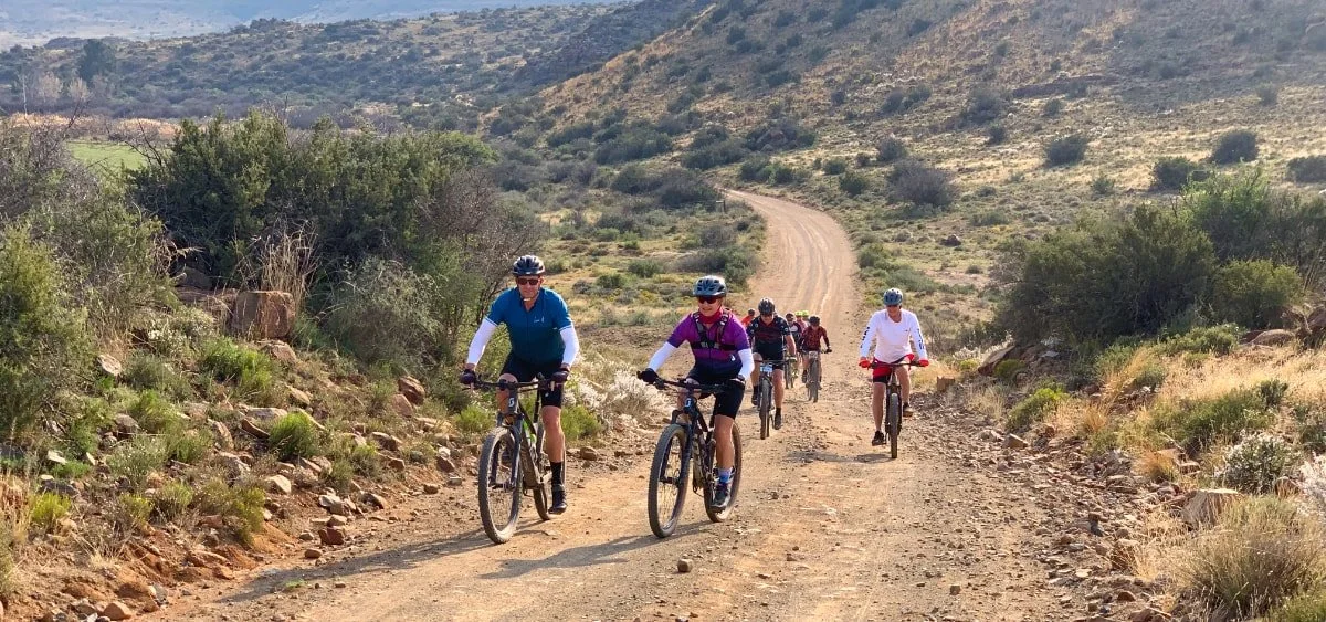 Group of people riding mountain bikes on a dirt trail in a desert landscape with sparse bushes and hills.
