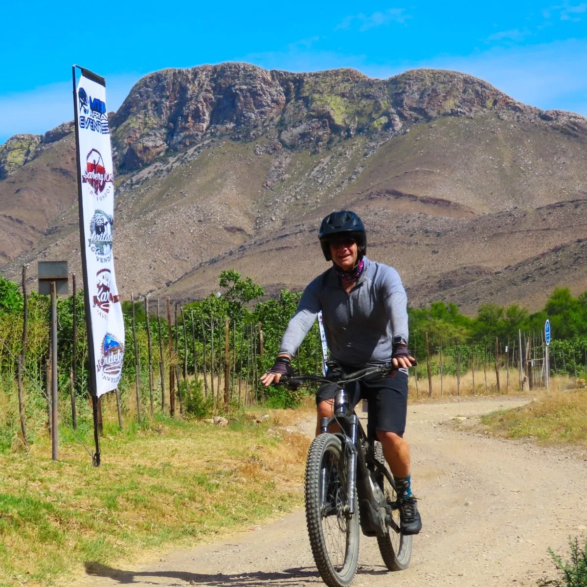 A person riding a mountain bike on a dirt trail with mountains in the background and a blue sky.