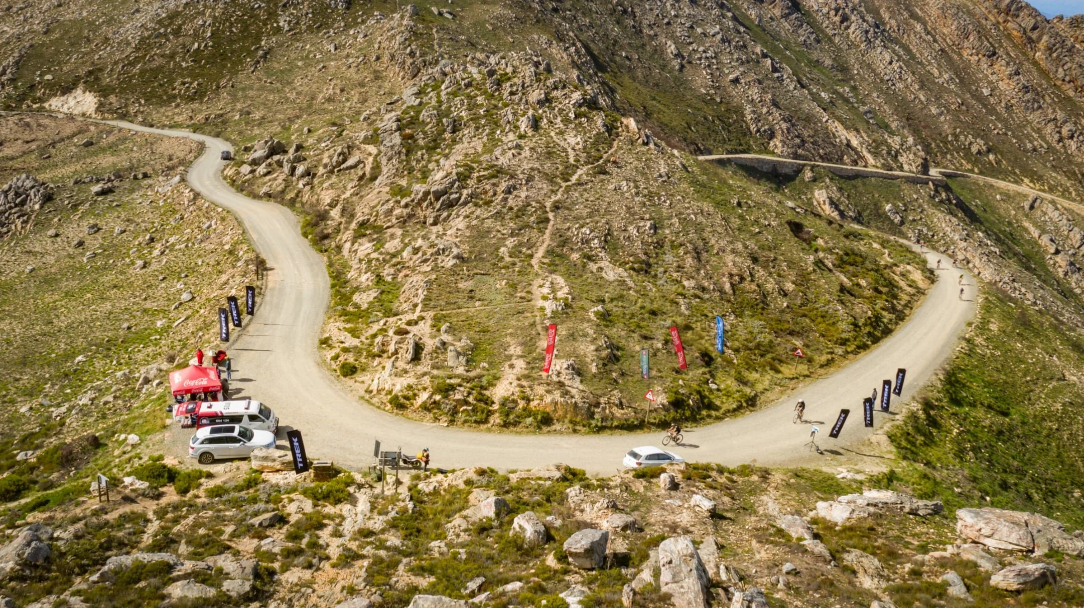 A winding mountain road with cars, cyclists, and banners for a cycling event or race.