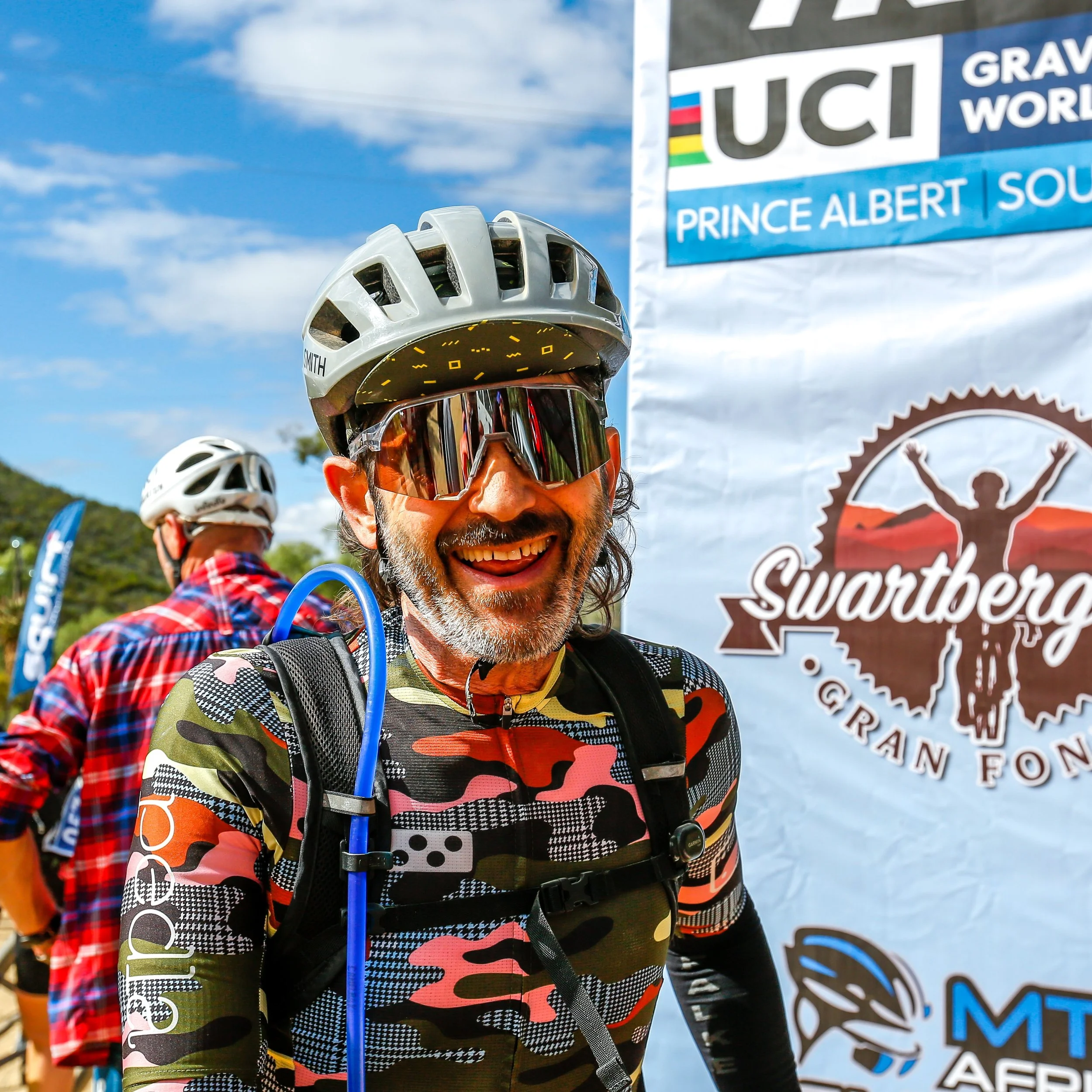 Smiling man in cycling gear, helmet, and sunglasses at a cycling event with a banner in the background.