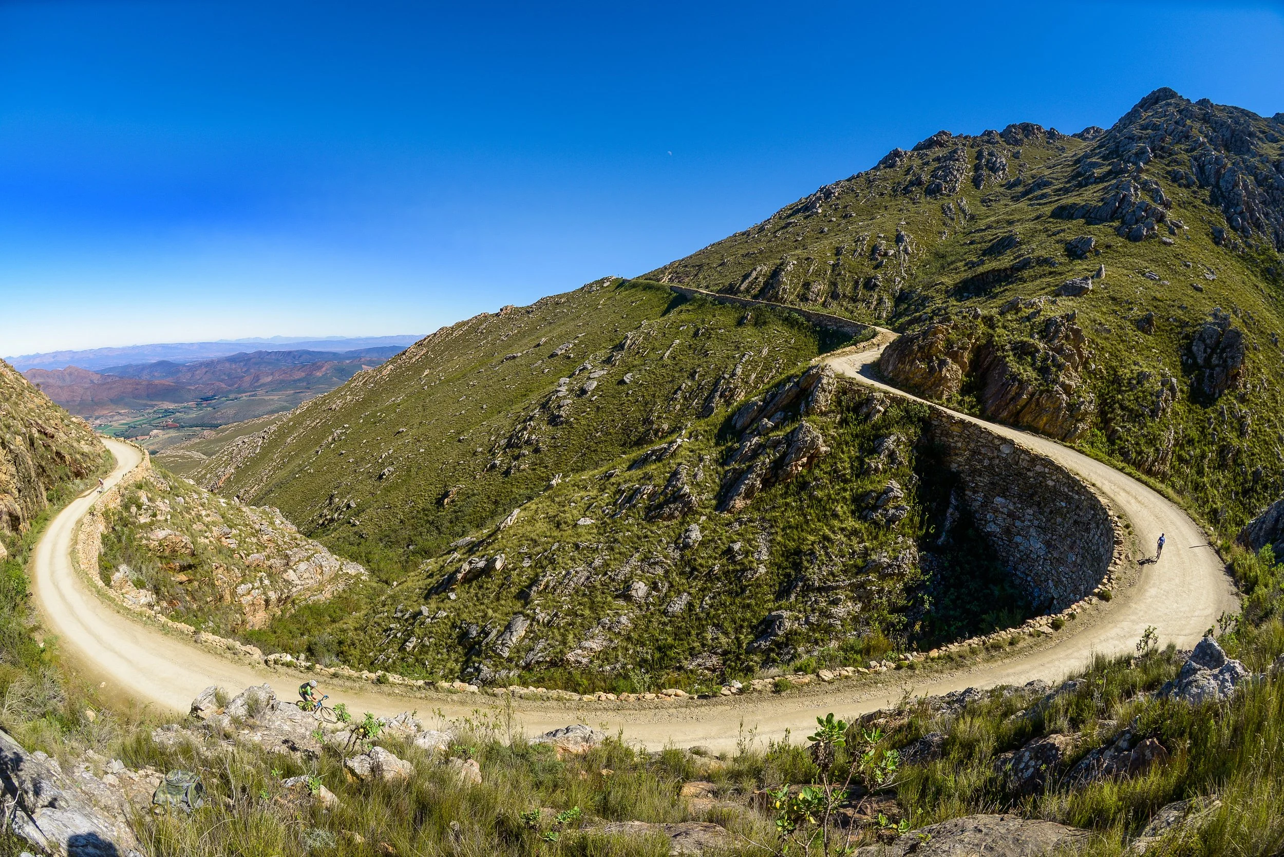 A winding dirt road ascending a green, rocky mountain under a clear blue sky, with a cyclist riding along.