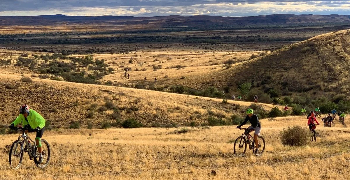 Group of cyclists riding on a dirt trail through a hilly, grassy landscape with sparse trees and distant mountains under a cloudy sky.