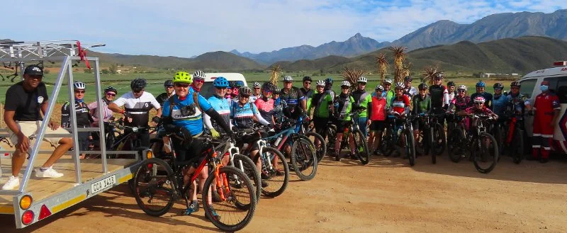 Group of cyclists with mountain bikes and helmets, some with safety gear, gathered outdoors with mountains in the background.