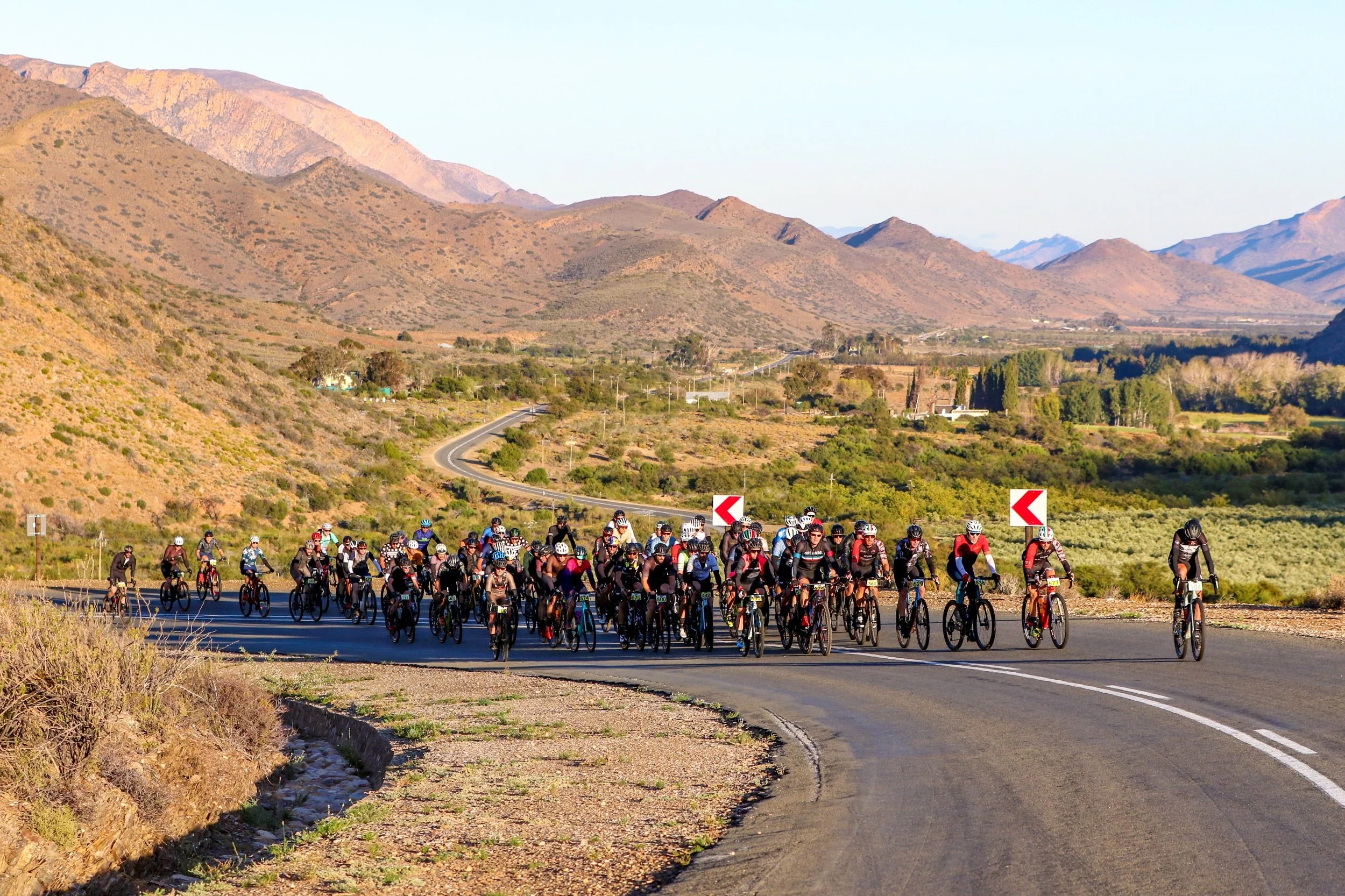 Group of cyclists riding on a winding mountain road with mountains and hills in the background.