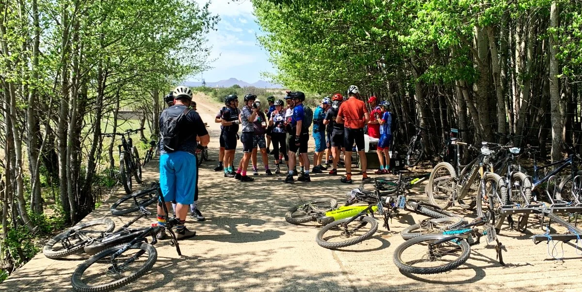 Group of cyclists gathered on a dirt trail with their bikes parked nearby, surrounded by trees and a fence, on a sunny day.