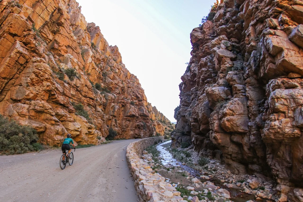 A cyclist riding a mountain bike on a dirt trail through a narrow canyon with steep rocky walls and a small stream running alongside.