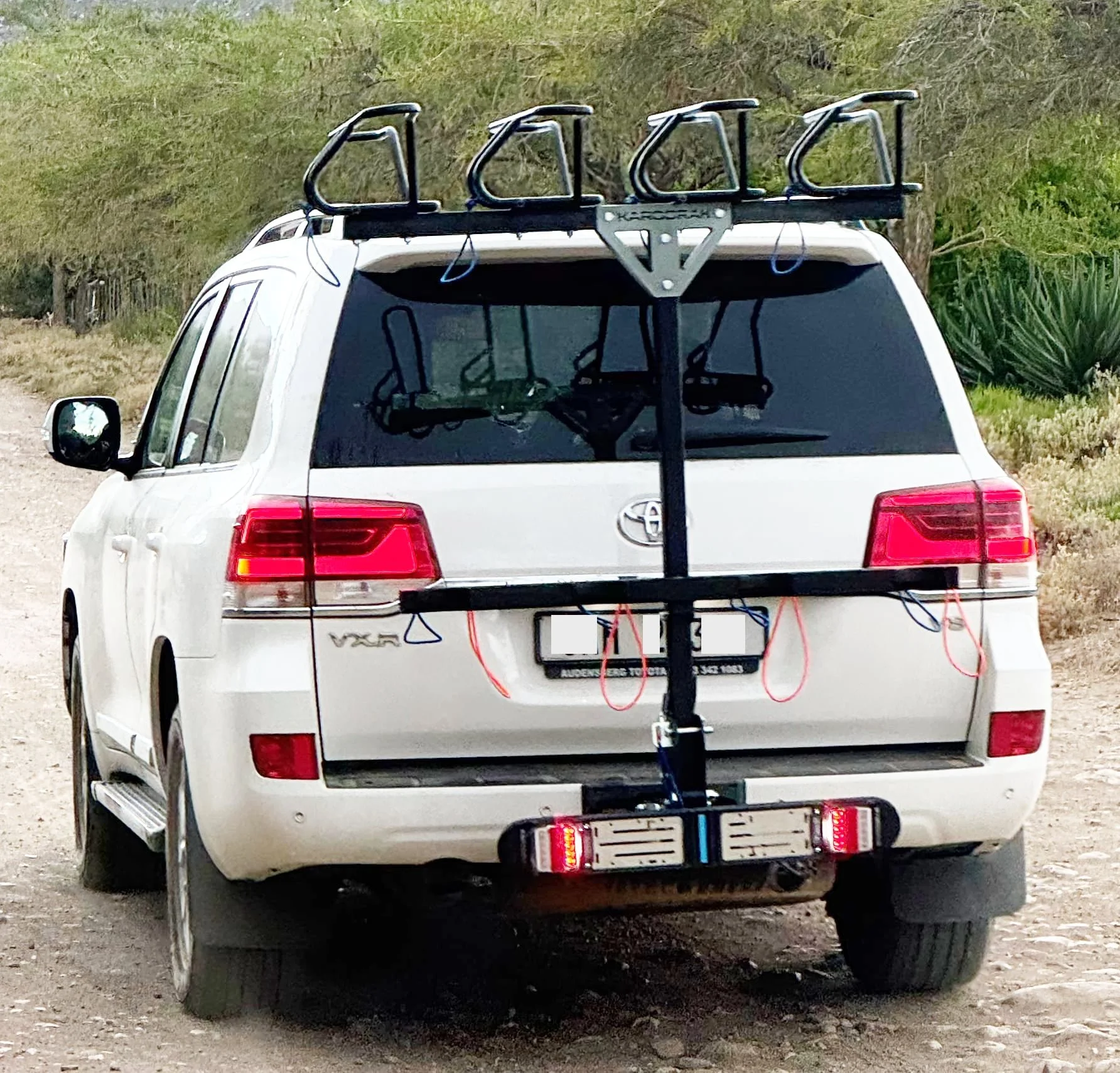 White SUV with bike rack attached to the rear, on a dirt path with greenery and desert plants in the background.