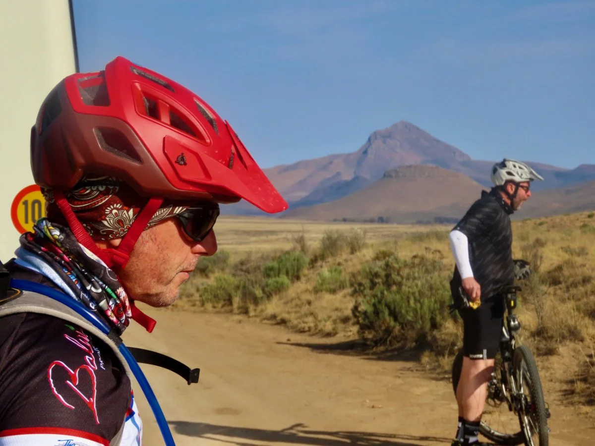 Two cyclists with helmets and sunglasses standing on a dirt trail in a desert landscape with mountains in the background.