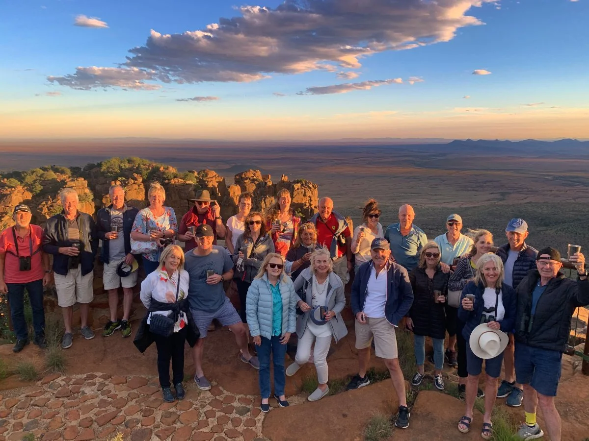 Group of people standing together on a rocky overlook at sunset, with a scenic landscape of rock formations, open plains, and mountains in the background.