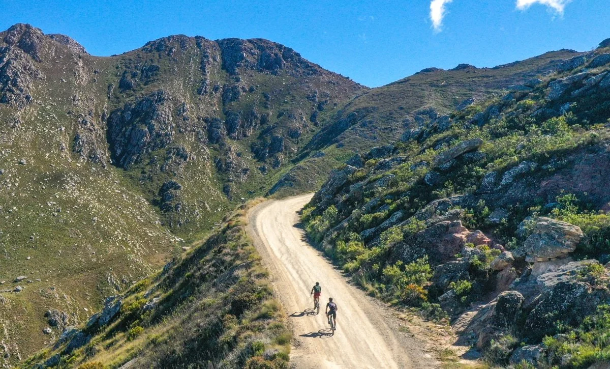 Two cyclists riding on a dirt trail through a mountainous landscape with green vegetation, rocky hills, and a clear blue sky.