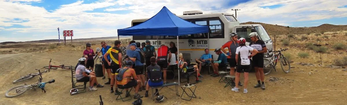 Group of cyclists resting and socializing under a blue canopy tent on a dirt landscape, with bicycles leaning against the ground and a support vehicle parked behind.