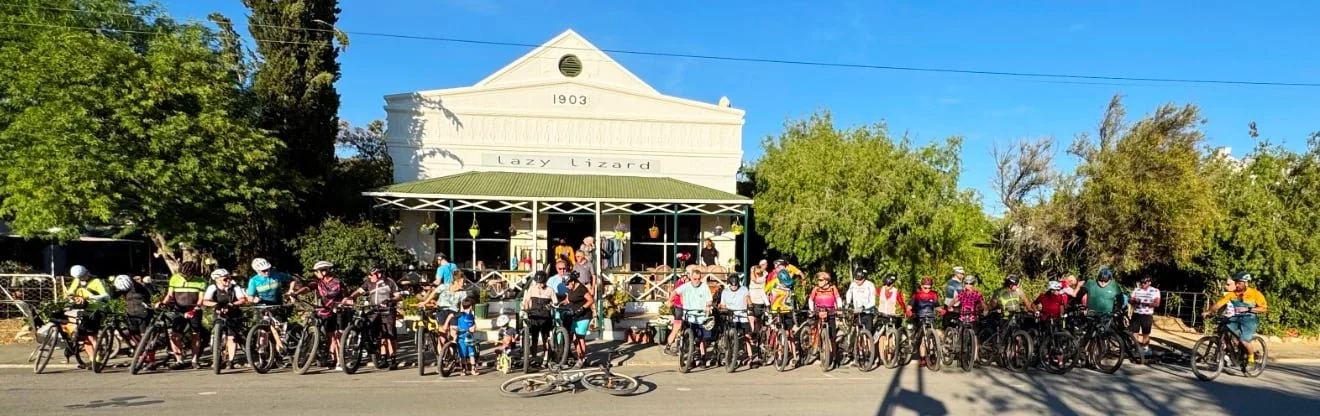 A group of cyclists gathering in front of a white building with a sign that reads 'Lazy Lizard' and the year 1903. The cyclists are lined up along the sidewalk next to their bikes, and some people are standing or sitting nearby. Trees and bushes are 