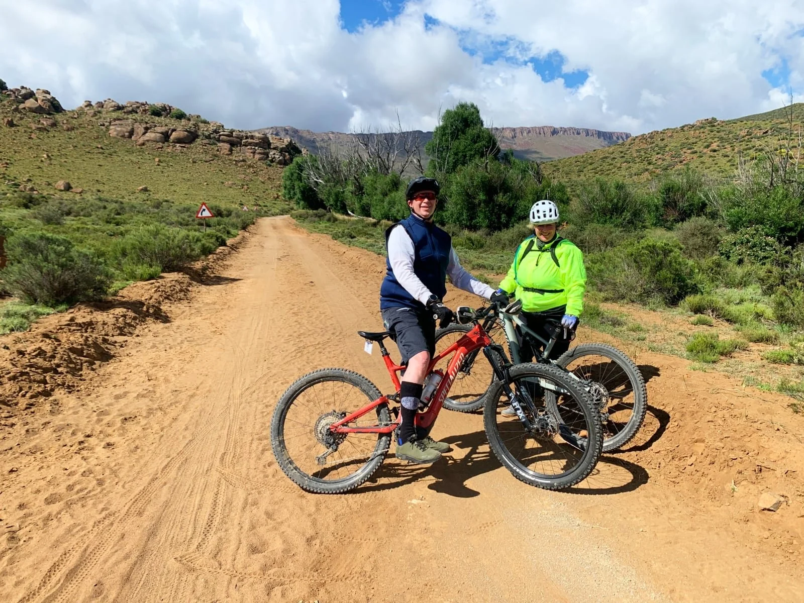 Two people in cycling gear standing with mountain bikes on a dirt trail in a hilly, green landscape under partly cloudy skies.