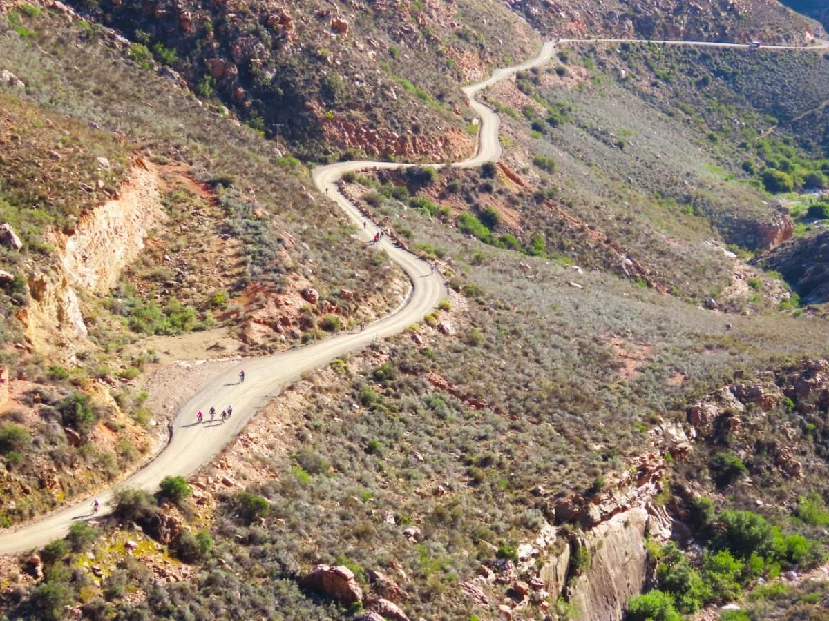 A winding dirt road through a hilly, desert-like terrain with sparse vegetation, and a group of hikers walking along the trail.