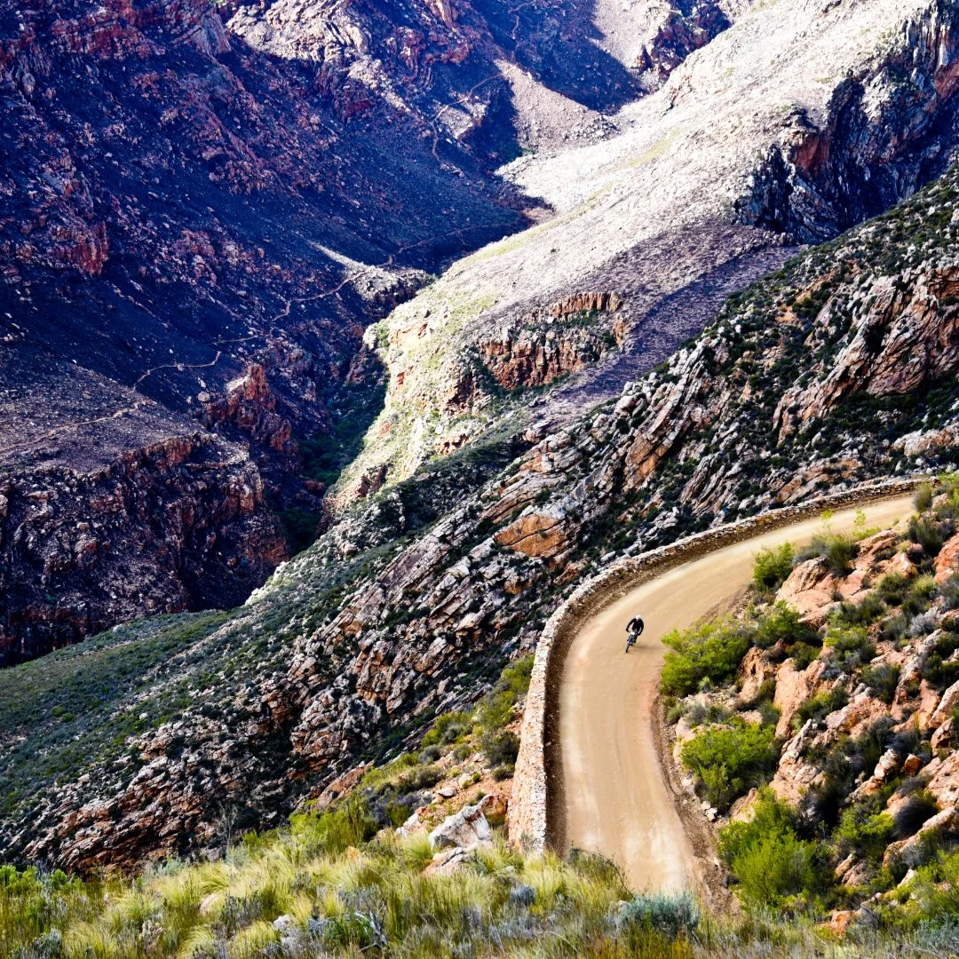 A cyclist riding along a dirt road that winds through a rugged canyon with steep rocky cliffs and sparse vegetation.