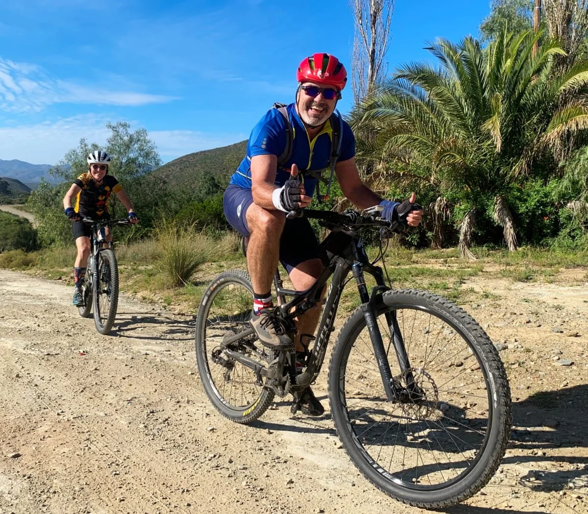 Two people riding mountain bikes on a dirt trail in a sunny, mountainous area with trees and blue sky.