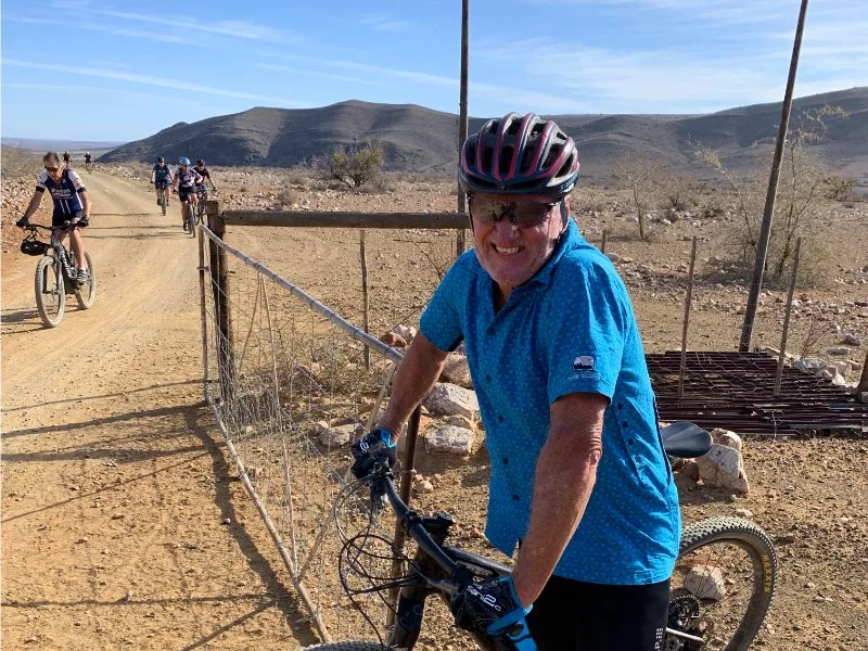 A man in a blue shirt and helmet smiling while riding a mountain bike on a dirt trail in a desert landscape, with other cyclists in the background.