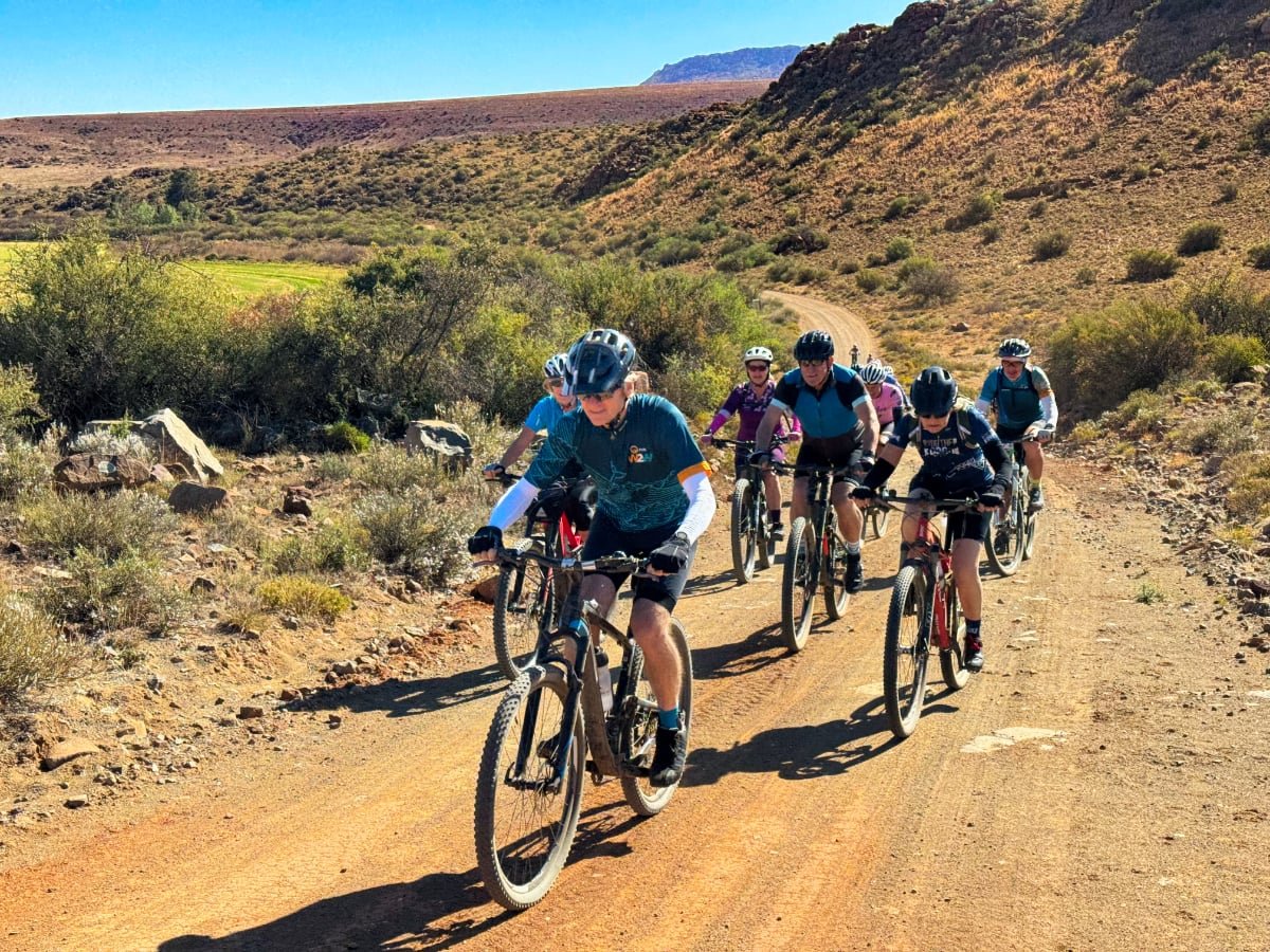 Group of seven people riding mountain bikes on a dirt trail through a desert-like landscape with bushes and hills in the background.