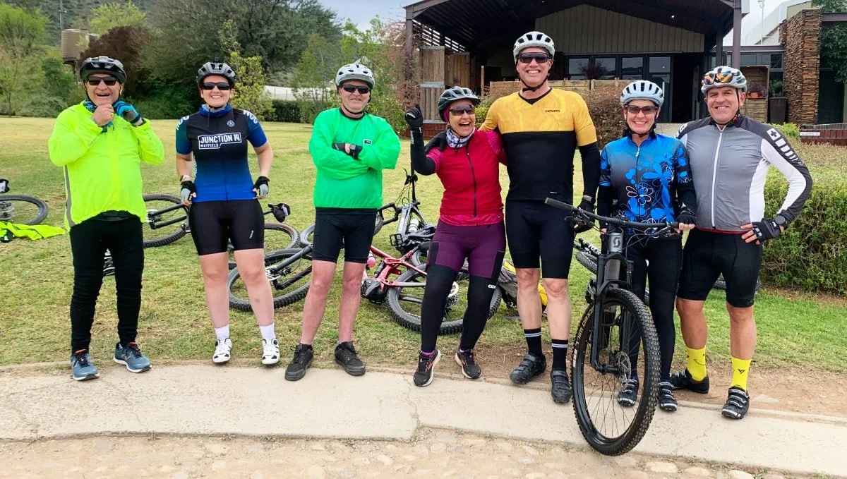 Group of eight cyclists in colorful gear, standing outdoors with their bikes, smiling, and posing together.