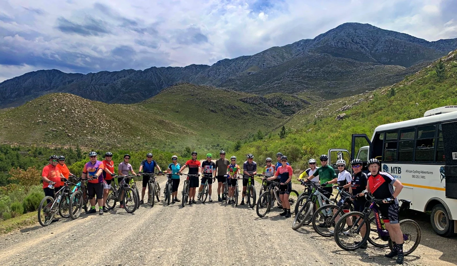 Group of people with bicycles on a dirt trail in a mountainous area with green vegetation and cloudy sky.