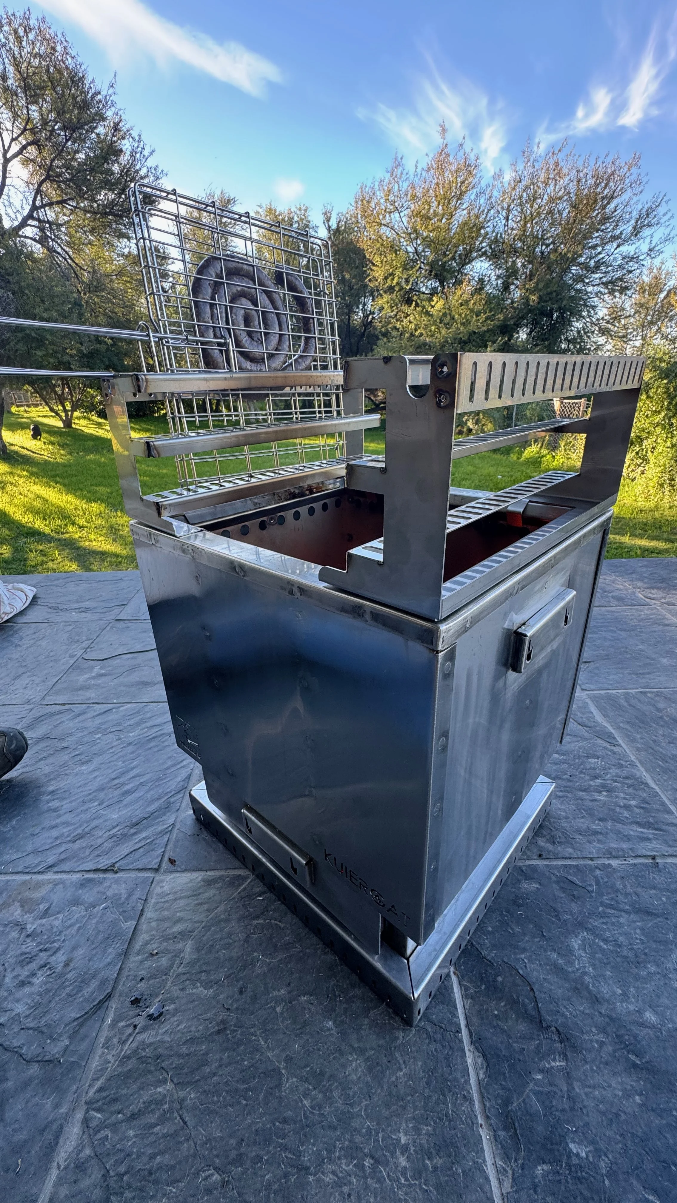 A stainless steel outdoor grill with a spiral sausage on a cooking rack, set on a stone patio with trees and blue sky in the background.