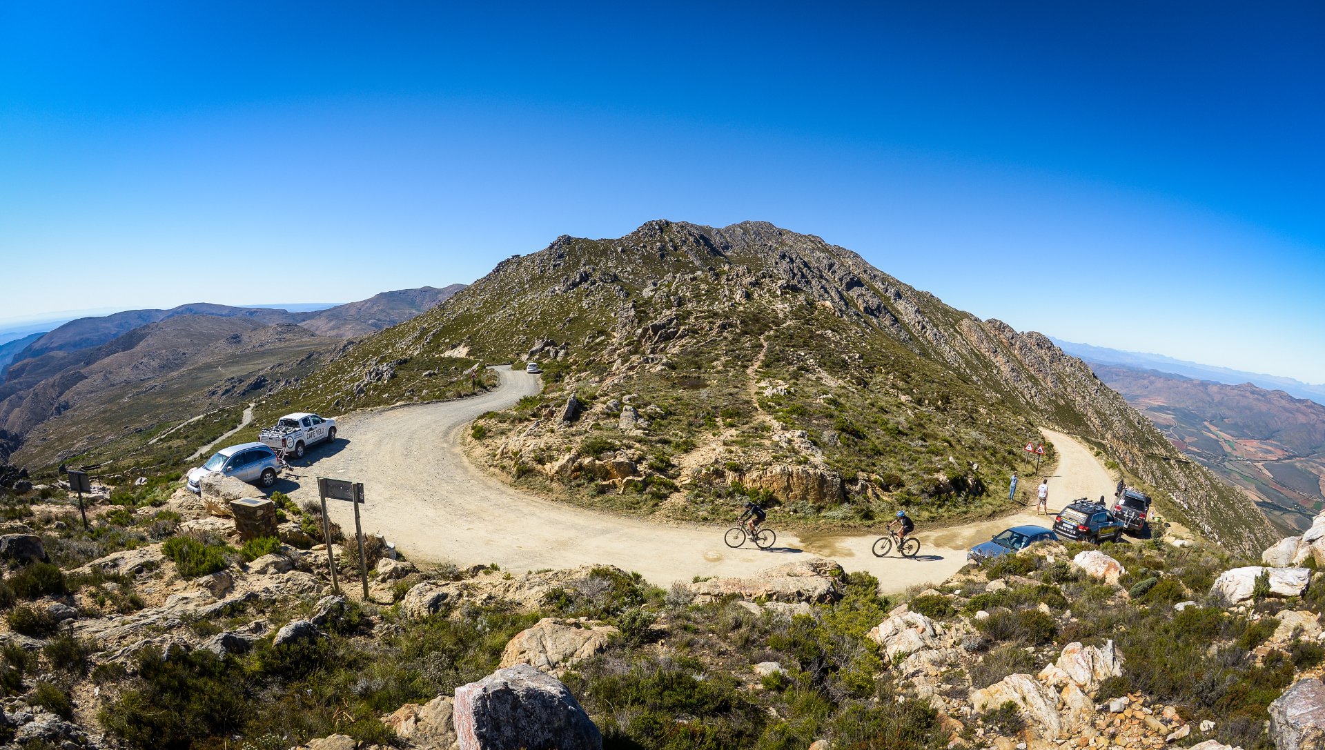 Scenic mountain view with a winding dirt road, parked cars, cyclists riding uphill, and a person standing near traffic signs under a clear blue sky.